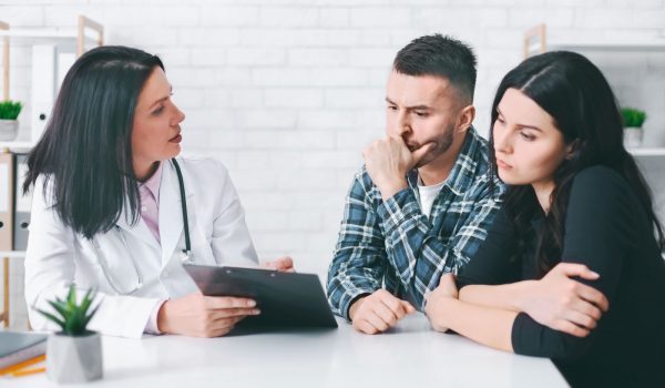 A doctor sits at a table with a young couple. The doctor is holding a clipboard and appears to be talking with the couple. The couple looks concerned and listens intently to the doctors advice.
