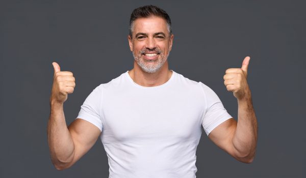 Happy fit sporty older man coach, middle aged personal trainer wearing white t-shirt showing like thumbs up standing isolated on gray background motivating for good results giving recommendation.