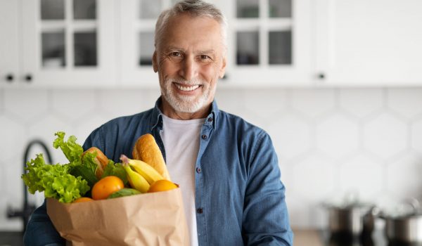 Happy Retirement. Smiling Senior Man Posing With Grocery Bag In Kitchen Interior, Cheerful Elderly Gentleman Holding Pack With Fresh Vegetables And Fruits And Looking At Camera, Copy Space
