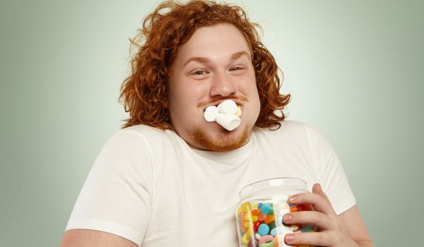 Headshot of funny greedy fat European man with ginger curly hair posing at studio, holding glass jar of sweets, with his mouth full of marshmallow, looking at camera with joyful happy expression