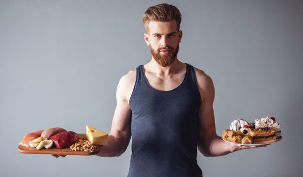 Handsome young bearded sportsman is holding a wooden tray with healthy food and a plate with cakes, on gray background
