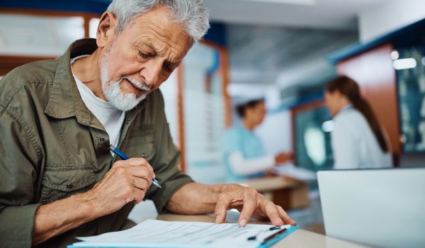 Senior man writing his data in medical report at reception desk in the hospital.