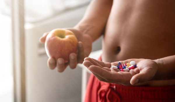 Close up of confused, puzzled man holding red fresh apple in one hand and pill, vitamins in another. He is trying to decide which choice is the best one. Horizontal shot